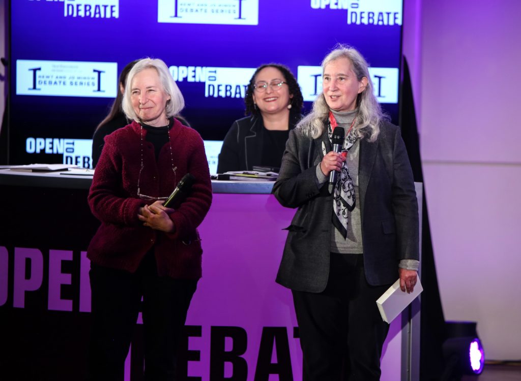 Martha and Nell Minow speak on the debate series stage as debate participants look on in background.