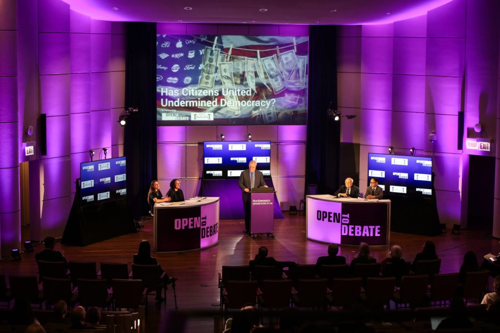 A stage backlit in purple, with a moderator in the center and two debate participants on either side of the moderator. The powerpoint text at the back of the stage reads "Has Citizens United Undermined Democracy?"