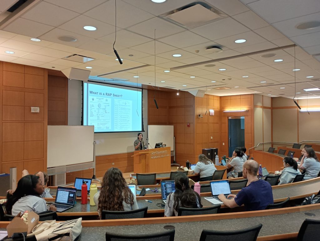 Image of students seated in a lecture hall while a speaker stands at the podium.